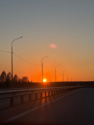 The image depicts a sunset over a road in Ivanovka, Russia. A guardrail separates the road from a line of trees. Streetlights line the side of the road, with the sun appearing to be positioned behind one of them. The sky transitions from a dark blue-gray at the top to an orange-yellow near the horizon. A vehicle is visible further down the road.