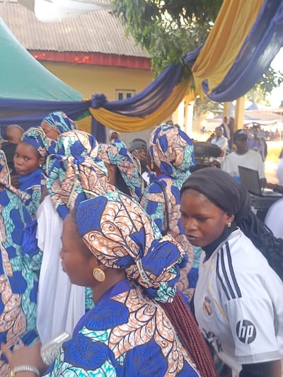 A group of women dressed in matching blue and brown patterned outfits and headwraps fill the frame. A woman in the foreground wears gold disc earrings and a headwrap that reads "WAN WAX WAX". Beside her, a woman with a black headscarf and a white soccer jersey with black stripes looks toward the right. In the background, a yellow and blue draped cloth hangs from a building. Other people are visible in the distance.