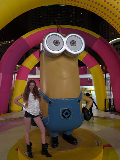 A young woman poses next to a large Minion sculpture in Shanghai, China. The Minion is surrounded by colorful, inflatable arches. The woman wears a white shirt, dark shorts, and black boots. Buildings are visible in the background. The ground has a colorful, swirled pattern.