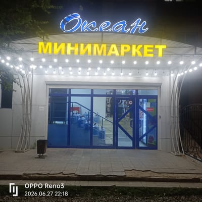 An exterior shot at night captures a brightly lit minimarket with a neon sign reading "Okean" in blue and yellow. Below the "Okean" sign, in large yellow letters, is the word "МИНИМАРКЕТ" (Minimarket). The entrance of the minimarket features double blue glass doors. Inside, a staircase is visible, with several large blue water bottles stacked on its steps. The minimarket is adorned with strings of small, round lights that hang from the awning, creating a festive atmosphere. The pavement in front of the store is made of interlocking bricks, some of which are broken or displaced. A dark-colored trash receptacle stands to the left of the entrance. The scene is located in Atyrau, Kazakhstan, as indicated by the timestamp "2026.06.27 22:18" and the "OPPO Reno3" branding, which are common on images taken in that region.