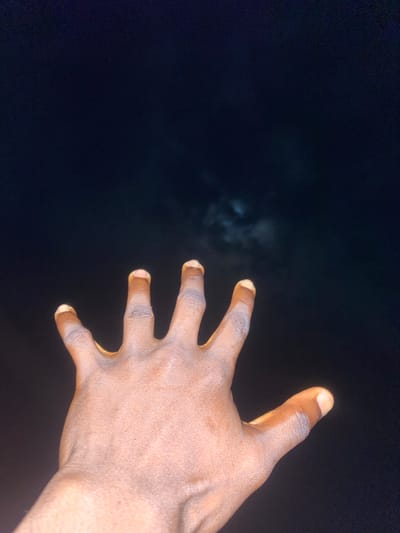 A close-up, low-angle shot captures an individual's left hand, palm facing upwards, reaching towards a dimly lit, cloudy night sky. The hand is dark-skinned with visible veins and appears to be the primary subject. The background is a dark, indistinct sky with faint, wispy clouds partially obscuring what might be a distant light source, possibly the moon. The scene suggests a solitary action or moment of contemplation in an outdoor environment. The location is Tavo, Nigeria.