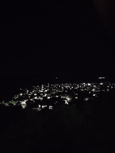 The image shows an aerial view of a town at night, with numerous lights illuminating the residential areas. The lights appear to be from houses and streetlamps, suggesting a populated area. The foreground is dark, with silhouetted vegetation, indicating a vantage point from a higher elevation overlooking the town. The sky is completely dark, with no visible celestial bodies, emphasizing the nighttime setting. The overall impression is a peaceful, albeit somewhat distant, view of a town illuminated against the darkness. This scene is consistent with the town of Kostenets, Bulgaria, known for its picturesque location and residential areas that would be illuminated at night.