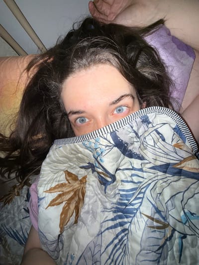 A person with striking blue eyes peeks out from under a patterned quilt. The quilt features large leaves in shades of blue and brown on an off-white background, with a black and white striped edge. Dark, wavy hair frames the face, with hints of gray visible near the forehead. The person appears to be lying down, as evidenced by the glimpse of a pillow and bed frame in the background. The perspective is slightly skewed, suggesting the photo was taken from above and at an angle.