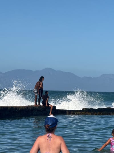 The image depicts a seaside scene in Muizenberg, South Africa. A concrete structure, likely a sea wall or tidal pool edge, runs vertically through the center. Waves are crashing against the wall, creating white foam. Three men stand on the landward side of the structure. Two appear to be shirtless, while the third is wearing what appears to be a black shirt and shorts. Another person is on the water side of the structure and wearing a pink t-shirt. A woman, back to the camera, is closer to the viewer, wearing a blue and white hat and a bikini top. Beyond the wall, the ocean stretches to the horizon, and mountains are visible in the background under a clear blue sky. The image is slightly rotated.