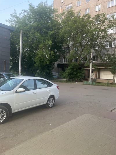 The image depicts a street scene in the city of Shebunino, Russia, on a cloudy day. In the foreground, a white sedan is parked on the asphalt. To the right, a paved pedestrian walkway is visible. Behind the car, a multi-story brick building with numerous windows stands tall. Lush green trees line the building's facade, obscuring parts of it. A utility pole with wires runs vertically through the scene. On the ground floor of the building, there appears to be a commercial establishment with a sign that reads "ПАЙРИМААЛПЕРСКАЯ." A set of stairs leads up to the entrance, and a small, green decorative fence is placed in front of it. Another dark-colored building is partially visible on the left side of the image.