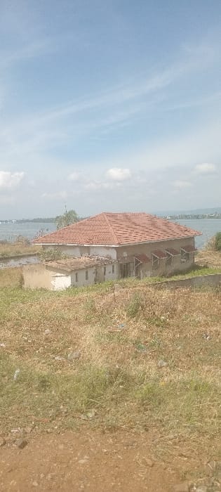 The image displays a static outdoor scene situated along the shores of Lake Victoria in Kisumu, Kenya. A single-story building with light-colored, weathered walls and a reddish-brown tiled hip roof is centrally located in the midground. An attached smaller, lower annex with a flat roof is positioned to the left of the main structure. The primary building features multiple windows with external sunshade-like covers visible on its right facade. Both structures exhibit signs of disrepair and weathering. Directly behind the buildings lies a narrow body of water, likely a flooded margin or shallow area, which precedes the open expanse of Lake Victoria. Distant landmasses are visible across the lake under a clear, light blue sky with scattered white, wispy clouds. The foreground consists of dry, sparse yellowish-brown vegetation and scattered debris on unmaintained ground, with a visible dirt path at the bottom. No individuals or actions are present in the scene.