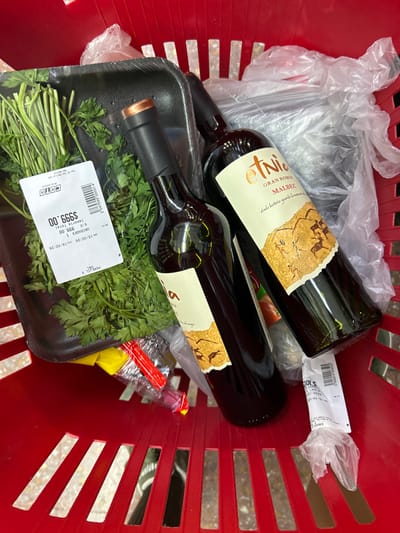 A red shopping basket contains two bottles of "Tinogasta Gran Roble Malbec" wine. One bottle lies diagonally across the top. A package of parsley in a black foam tray sits at the bottom, featuring a COTO price tag of $999.00, dated 15/02/26. Clear plastic bags are present, obscuring other items. A receipt with additional information is partially visible. The perspective is an overhead shot of the basket's contents.