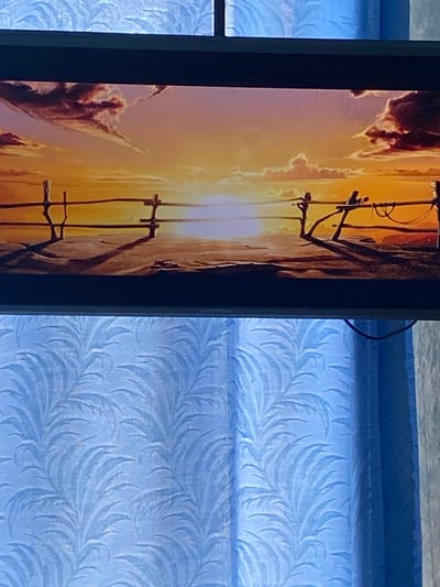 The image shows a television screen displaying a sunset over a body of water, with a wooden fence in the foreground. The screen is positioned in front of blue curtains with a leaf pattern. The scene on the screen is an animated illustration of a sunset with vibrant orange and yellow hues. The sun is low on the horizon, casting long shadows from the fence posts. The water reflects the colors of the sky. The curtains behind the television are a light blue and have a repeating pattern of stylized leaves. The overall impression is of a tranquil, natural landscape depicted on a modern electronic device. The context of Mamontovo, Russia is not visually evident in the image itself.