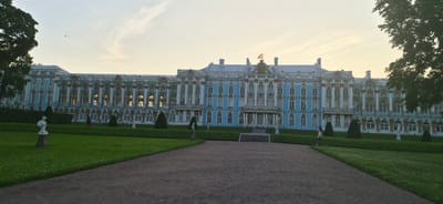 The image shows the Catherine Palace in Pushkin, Russia, a grand blue and white baroque-style building with ornate golden detailing. The palace is set against a pale sky with faint clouds, and the lighting suggests it is either early morning or late afternoon. In the foreground, a paved path leads towards the palace, bordered by a lush green lawn. Several conical evergreen trees are strategically placed along the front of the palace grounds. On the lawn, there are at least two white classical statues on pedestals.
Several people are visible: two individuals, possibly tourists, are walking on the lawn near the palace. Another person, dressed in white, stands facing the palace on the right side of the frame. A few other figures are indistinctly seen near the palace entrance. A single bird is captured in flight in the sky above the palace. The overall scene conveys a sense of historical grandeur and tranquility.