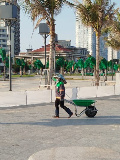 A worker wearing protective clothing is walking and pulling a green wheelbarrow across a paved area in Ho Chi Minh City, Vietnam. The worker is wearing a wide-brimmed hat, face covering, dark clothing, a gray and orange striped jacket, and boots. Rows of trees covered in green mesh line the background, and buildings are visible in the distance. A temporary white barrier is present. The day appears sunny.