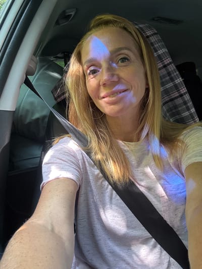 A woman is sitting in the passenger seat of a car, wearing a pink shirt and a seatbelt. There is a plaid blanket behind her. She has blonde hair and is smiling at the camera. The car appears to be stationary. The image was taken in Portovoe, Russia.