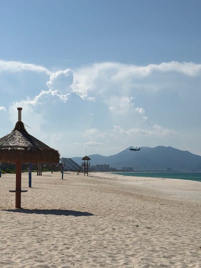 A wide, sandy beach is depicted under a bright, blue sky with scattered cumulus clouds. In the foreground, a thatched umbrella casts a shadow on the sand. A wooden lifeguard tower stands further back on the beach, with buildings and mountains visible in the distance. An airplane is flying in the sky, with its landing gear down. The ocean, a strip of turquoise, is visible in the background. This scene is characteristic of a beach resort area.