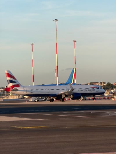 The image depicts an airport tarmac scene at Fiumicino, Italy, under a clear, pale blue sky with soft, directional lighting suggesting late afternoon or early morning.

In the foreground, a British Airways commercial aircraft is parked, identifiable by its white fuselage, blue underbelly, and the Union Flag livery on its vertical stabilizer. The registration "G-NEOP" is visible on the rear fuselage. A mobile passenger stair is positioned against an aft door on the port side of the aircraft. Engine cowlings are dark blue. Orange traffic cones and various ground support equipment, including what appears to be a service vehicle, are visible around the aircraft.

Behind the British Airways aircraft's wing and tail, a second commercial airplane is partially visible. Its vertical stabilizer is distinctly painted with vertical stripes of green, white, and red, characteristic of an Italian airline. This aircraft's fuselage appears to be a light color.

The background features several tall, slender poles with alternating red and white horizontal stripes, characteristic of airport lighting or navigational aid towers. One such pole is prominently positioned between the two aircraft, and two others are visible further back on the right and left. Distant structures and buildings, typical of an airport perimeter, are discernible on the horizon. The tarmac in the foreground displays yellow and white painted markings. No individuals are clearly discernible.