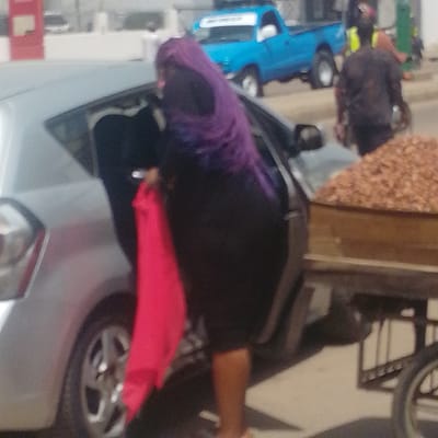 In Arawa, Nigeria, a scene unfolds involving a silver car and a food cart. A person in a black garment and a purple headscarf is seen near the open driver's side of the car, holding a bright pink cloth. In the background, a blue pickup truck is parked. To the right, a food cart is visible, laden with what appears to be a large quantity of nuts or similar food items. Another individual, partially obscured and wearing dark clothing, is also present near the cart. The environment appears to be outdoors, possibly a street or market area, with a blurred background suggesting buildings and possibly other vehicles.