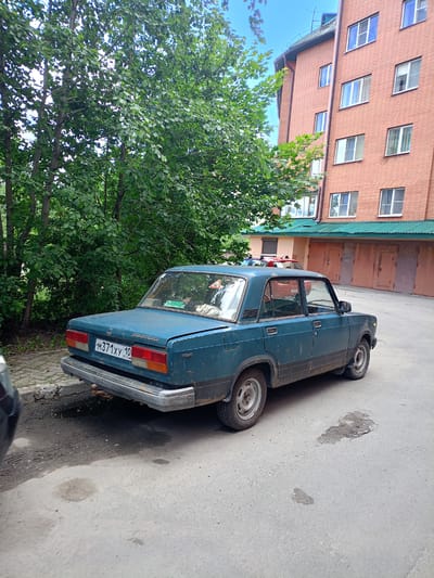 The image shows a dark teal Lada sedan parked on a paved surface next to a sidewalk and a line of trees. The car is viewed from the rear passenger side. The license plate reads "M371xy 10". Behind the car, a reddish-orange brick building with multiple windows and a green roof is visible. The building appears to be a multi-story apartment complex or similar structure with garages on the ground floor. The sky is blue with some white clouds. The overall scene suggests a residential area in Petrozavodsk, Russia, likely in a courtyard or parking area adjacent to an apartment building. There are no people visible in the image.