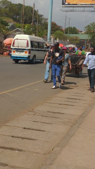 This image depicts a daytime street scene in Moshi, Tanzania. The scene includes a paved asphalt road with a faded yellow line marking the shoulder, adjacent to a concrete sidewalk featuring multiple linear drainage channels.
On the road, a white minibus (known locally as a Dala Dala) with dark horizontal stripes, a blue rear bumper, and yellow/red chevron reflective tape is either parked or moving slowly. Its rear license plate begins with "T 908". To the left of the road, several street vendor stalls with orange and red umbrellas are visible.
A group of four pedestrians is walking towards the right side of the frame, along the edge of the road, next to the sidewalk. From left to right:
1. A person wearing a dark top and light blue jeans, with a white cap, carrying a bag over the left shoulder.
2. A person wearing a black t-shirt with a light circular emblem, blue jeans, a black cap, and white sandals, carrying a dark blue bag.
3. A person wearing a green t-shirt, patterned trousers, and a pink cap.
4. Further right, a male individual wearing a light blue patterned shirt is partially visible.
To the right of the walking group, a male individual in a green shirt is pulling a wooden handcart filled with a dark, granular material.
In the background, there are various trees, residential and commercial buildings (some with green roofs), utility poles (one light blue, others brown), and a large billboard structure displaying orange and yellow graphics at the top right of the frame. The sky is partially visible and appears clear.