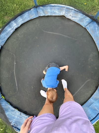 A high-angle shot depicts a child in a blue shirt and gray shorts crawling on a black trampoline. An adult wearing a lavender outfit and white socks is also on the trampoline. The trampoline has a blue padded safety edge. The background shows grass and a hint of trees, suggesting an outdoor setting. The location in Ферма, Russia is not verifiable from the image.