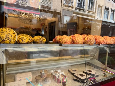 The image captures a shop window display in Lyon, France. A variety of sweets, including giant cookies with chocolate chips and pink praline brioches are arranged on the upper shelf. Below, there are tubs of ice cream, boxes of Florentin cookies, and Les Ballotins chocolate boxes. A reflective window provides a distorted view of the surrounding buildings, which include apartments and commercial storefronts. The reflection shows decorative architectural details, window shutters, and awnings. There is a chandelier visible in the background, suggesting a luxurious interior. Text on the storefront includes "la grande" above the cookies and “Pralus Meilleur” on the edge of the brioches' wrapper. “Francois Pralus” is written on the various containers and boxes.