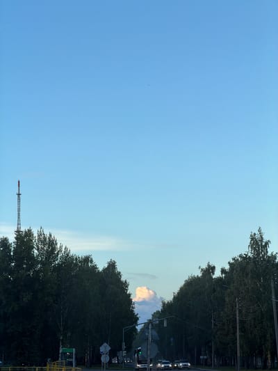 This is a vertical, daytime, outdoor shot of a clear blue sky with a few wispy clouds. On the right side of the frame, a dense line of dark green trees runs along the edge of a road. Several vehicles, including white vans or SUVs, are visible on the road, suggesting traffic. There are also streetlights and traffic signals. In the distance, a tall, lattice-style radio tower with red and white markings can be seen against the sky. The overall impression is of a suburban or roadside scene on a bright, clear day. The image was taken in Nizhnekamsk, Russia.