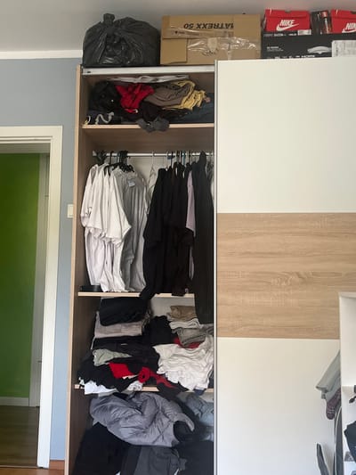 An indoor scene captures a wardrobe with sliding doors, one white and the other with a light wood grain finish, currently open to reveal its contents. The lower section of the wardrobe is filled with various items of clothing, some neatly folded white shirts on hangers, while others are piled in shades of black, red, grey, and beige. A large black trash bag rests on the bottom shelf to the left. Above the wardrobe, stacked cardboard boxes are visible, including several Nike shoe boxes, a box labeled "MATREXX 50," and another for a "ASUS GAMING MONITOR" with details like "PRIME B450M-K," "144," and "HDMI." A box for "LEGO TECHNIC" is also present. The walls surrounding the wardrobe are painted white, with a section below the wardrobe in a light blue color. A white door frame is visible on the lower right, revealing a bright green interior space beyond. The image depicts a storage area, possibly in a bedroom, with a mix of organized and unorganized items. The location context is Pokrovnik, Bulgaria.