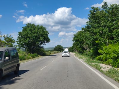 The image depicts a view from a moving vehicle on a paved road in Yasen, Bulgaria. The road stretches ahead, flanked by lush green trees on both sides, creating a natural tunnel effect. The sky is a vibrant blue, dotted with fluffy white clouds, suggesting a pleasant, sunny day.
In the foreground on the left, a silver van is partially visible, indicating the perspective of the viewer. Further down the road, a white car is driving away from the camera, its taillights visible. In the distance, there are rolling hills and more trees, adding depth to the landscape. The road itself has white lane markings, indicating it is a public thoroughfare.
The overall scene evokes a sense of travel and a peaceful journey through a rural or semi-rural area. The time of day appears to be midday or early afternoon due to the bright sunlight and the clear sky. There is no visible text in the image, other than potentially on the license plate of the white car, which is too blurry to read.