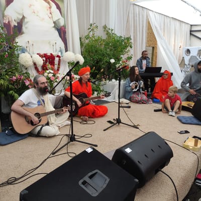 In Zhirnosovo, Russia, a group of people are gathered outdoors, possibly for a religious or cultural event. A large portrait of a person in traditional attire is displayed behind a table adorned with flowers and candles. Several individuals are seated on the ground, some playing musical instruments like guitars and a keyboard. One person wears an orange robe and turban, while others are dressed in casual clothing. A young child is also present, observing the scene. The environment appears to be a temporary setup with draped fabric and potted plants. Two large black speakers are positioned in the foreground, suggesting a musical performance or amplified speech is taking place. The overall atmosphere seems peaceful and communal.