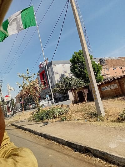 This image displays an urban street scene in Sharada, Nigeria, under a clear blue sky. A Nigerian flag, characterized by its green-white-green vertical stripes, flies prominently from a white flagpole in the upper-left quadrant. Multiple black overhead wires traverse the sky.
A light-colored concrete utility pole, marked with "101" in faded yellow paint, stands vertically on the right side of the frame. In the foreground, a paved concrete sidewalk is separated from a dirt road shoulder by a concrete curb. The shoulder contains dry grass and sparse vegetation. The lower-left corner of the image shows a partial view of an individual's arm and shoulder, dressed in light brown or khaki clothing.
In the midground, a dark-colored metal fence runs parallel to the road. Behind this fence, a multi-story building with a red and gray facade and white rectangular architectural elements is visible, featuring a prominent red "TATA" sign. To the right of this building, another structure with a reddish-brown tiled roof shows scaffolding on its side, indicating construction or renovation activity. A light-colored sedan is partially obscured behind the fencing. Scattered green trees and shrubs are present throughout the scene.