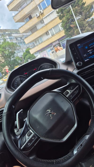 The image provides an interior view from a car, looking out through the windshield. The car's dashboard and steering wheel are prominently featured in the foreground, with the Peugeot logo visible on the steering wheel. On the dashboard, a digital screen displays "VITO..." and "37°C" along with radio information. Outside the car, a building with a yellowish facade and multiple windows and air conditioning units is visible, along with trees and a cloudy sky. In the background, another building structure can be seen. A yellow vehicle is partially visible to the left. The scene is consistent with an urban environment, specifically identified as the city of Sevlievo, Bulgaria, likely on a street. No people are clearly depicted. The image appears to be taken during daylight.