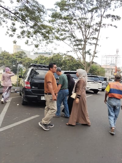 The image depicts an outdoor scene with several individuals and vehicles on an asphalt surface, possibly a parking lot or street. A black SUV is parked centrally, with its rear facing the camera. To the left of the SUV, a person in a light-colored patterned outfit and a purple headscarf walks away from the camera. In front of the SUV, a man in an orange polo shirt and khaki pants is walking, appearing to be in conversation with another person partially obscured behind him. To the right of the SUV, a woman in a beige hijab and dress walks towards the right side of the frame. Further to the right, a person wearing a yellow and orange vest over blue jeans walks away from the camera. Trees and buildings are visible in the background, suggesting an urban environment. The sky is overcast. The presence of trees and buildings does not definitively place the image in Bandung City, Indonesia, without further context.