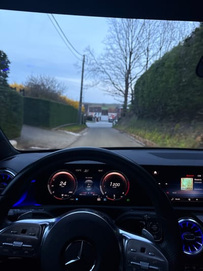 The image presents a first-person view from the driver's seat of a Mercedes-Benz vehicle in Tielrode, Belgium. The digital dashboard displays a speed of 24 km/h and an engine speed of approximately 1200 r/min. The outside temperature is 12.0°C, and the current time is 18:47. The infotainment screen indicates "MAABOLA" by "DOSAMIS" (Shamakova) is playing via Bluetooth/USB. The vehicle's interior features a multi-function steering wheel and blue-illuminated circular air vents.
Through the windshield, a narrow, paved road extends forward, appearing damp or wet. On both sides of the road, tall, dense green hedges line the immediate foreground. To the left, a utility pole with overhead wires is visible. Behind the left hedge, there are bushes with sparse yellow blossoms. Further down the road, bare deciduous trees are prominent, suggesting late autumn or winter. In the distance, a cluster of buildings with red-tiled roofs and red brick exteriors can be seen. The sky is overcast and light blue-grey.