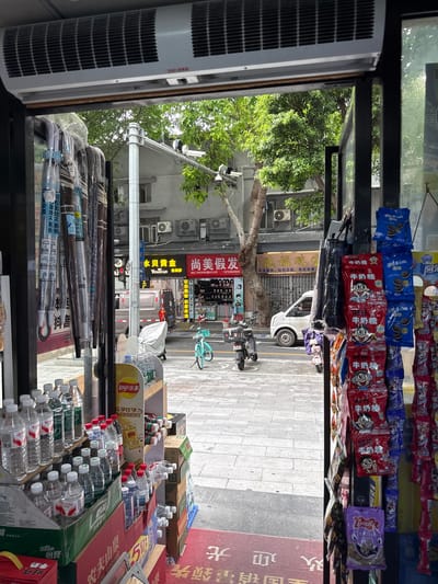 The image captures a street view through the open doorway of a store, likely in Liwan District, China. On the left, a rack displays several hanging umbrellas, mostly covered in plastic. To the right of the doorway, shelves are laden with bottled water, snacks, and other packaged goods.

Outside the store, a street is visible, paved with light-colored tiles. A white van is parked on the street. To the right, a turquoise bicycle is parked on the sidewalk, and a black electric scooter is also visible. In the background, buildings line the street, with some featuring air conditioning units mounted on their exterior walls. Trees with green foliage are scattered throughout the scene, providing shade. The overall atmosphere suggests a typical urban commercial area.