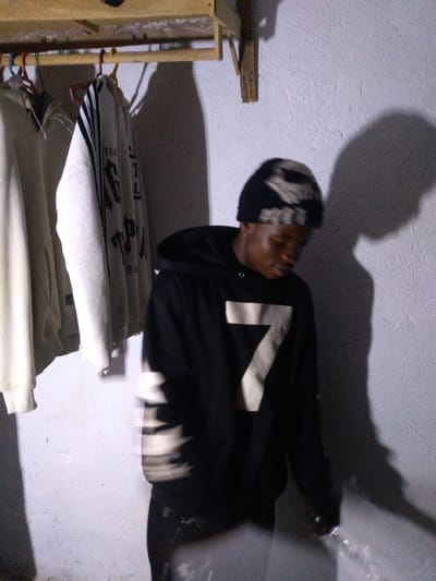 In an indoor setting in Unknown, Nigeria, a young Black male is captured in a medium shot, facing away from the camera with his head turned to his right. He is wearing a black beanie with a light-colored abstract pattern and a black hooded sweatshirt. The sweatshirt features a large, light-colored number "7" printed on the front. The background consists of a rough, textured white wall, casting a prominent shadow of the subject and the overhead clothing rack. To the left of the subject, several garments are hanging on a wooden rack suspended from the wall by wooden brackets. The clothing appears to be mostly hoodies or sweatshirts, in various neutral colors like beige and light gray, with some showing dark stripe patterns. The lighting in the scene is dim, creating a stark contrast between the illuminated subject and the shadowed background. The overall impression is that of a utilitarian, possibly unfinished, indoor space.