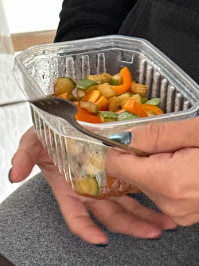 A person holds a clear plastic container filled with cooked vegetables, possibly leftovers. The vegetables appear to be diced green zucchini, orange bell peppers, and yellow squash. A metal spoon is inserted into the container, seemingly scooping out the vegetables. The person's hands are visible, with nail polish on their fingers. The background shows a black jacket, a gray surface, and a glimpse of a light-colored wall.