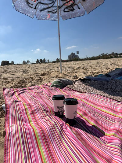 The image shows a beach scene in Ashkelon, Israel. A striped pink, white, green, and purple blanket is spread on the sand, partially covered by other blankets. Two white disposable cups with black lids, branded "Corona Extra", are placed on the blanket. An open beach umbrella, with a pattern of light blue squares and dark graffiti, stands behind the cups, casting a shadow. The background reveals a sandy beach, a line of trees and buildings in the distance, and a clear blue sky with a few white clouds. The overall impression is a relaxed, outdoor setting on a sunny day.