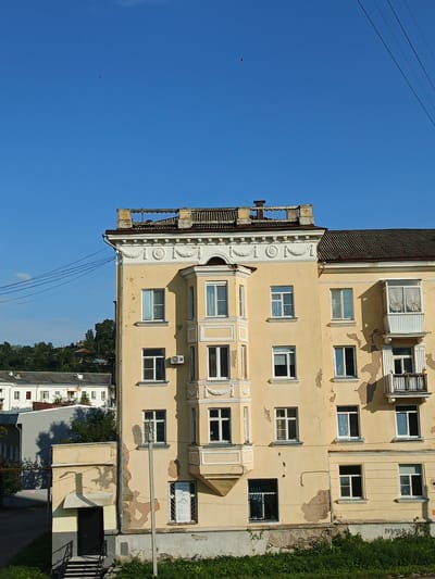 The image captures an exterior view of a light yellow, multi-story apartment building in Zlatoust, Russia. The building has a weathered facade with peeling paint and an ornate cornice. There are numerous windows of various sizes, some with decorative molding, and a few balconies, one of which has curtains visible. A protruding bay window structure extends from the front of the building. The roof appears to be covered in dark shingles, and there's a decorative railing along the top edge. In the distance, a lush green, tree-covered hillside with some houses is visible under a clear blue sky. Power lines crisscross the sky. At the lower left, a dark entrance door is set within a smaller attached structure with a gray, sloped awning and stairs leading up to it. The ground level of the building shows signs of disrepair. Green vegetation grows in front of the building. A single bird is visible in flight against the blue sky.