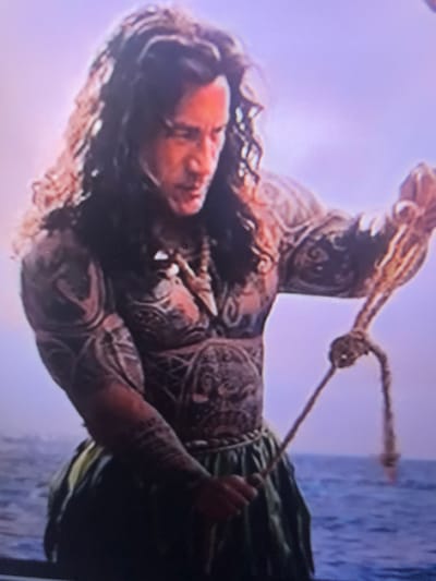 The image shows a close-up of a man with long, dark, curly hair and intricate tattoos covering his chest, arms, and shoulders. He is wearing a necklace with a pendant and a green skirt-like garment. He is holding a rope in his hands, seemingly in the process of tying or untying a knot. The background is a blurred seascape with a pale purple sky. There is no indication of the city of Solaize, France, in the image.