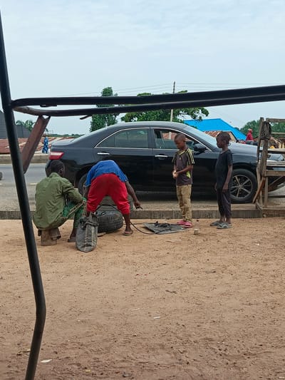 The image captures a street scene in what appears to be Sobon-Gida, Nigeria. A black sedan is parked on the side of a paved road. Two men are engaged in tire repair: one is sitting on a log stool, wearing a green shirt and patterned pants, while the other, in a blue shirt and red trousers, bends over a tire. A bag sits next to them. Two young boys stand nearby, observing the tire repair process. The ground is sandy. Buildings and a sky with partial cloud cover are visible in the background.