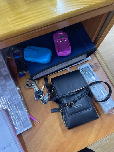 This media file shows an overhead view of a wooden drawer containing various items. The drawer appears to be part of a desk or cabinet, with visible wooden grain. The primary objects in the drawer are a black wallet with a gold zipper and a strap, a set of keys, a pink battery case, a blue electronic device, a remote control wrapped in plastic, and a pink pen resting on a notebook or planner. The wallet has "FOREVER YOUNG" and "Originally from Korea" printed on it. The remote control has buttons and some text indicating functions like "Mode," "Timer," and "Stop." The notebook is partially visible and appears to have a grid pattern on its pages, covered in plastic. The overall scene suggests a personal space, possibly a workspace or bedside table, where everyday items are kept. There are no people visible, and the lighting suggests it could be daytime, though no specific time or weather is indicated. The location context is Donskoye, Russia, which provides a geographical reference for the scene.