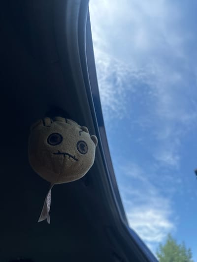 A plush toy resembling Groot is attached to the interior of a vehicle, possibly a car trunk or hatchback, viewed from inside the vehicle looking upwards. The plush toy, which is light brown with embroidered dark eyes and a mouth, hangs from a white tag. The background is a bright blue sky with scattered white clouds, and a hint of green foliage is visible in the lower right corner. The lighting suggests it is daytime. The location is not specified in the image or its context.