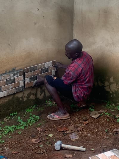 A bald Black man is kneeling on the ground and tiling a wall in Agban, Nigeria. He is wearing a red and black plaid shirt and dark pants, with flip-flops on his feet. He is placing a small, rectangular tile on a wall that is partially tiled with similar mosaic tiles. The wall appears to be made of rough concrete and is stained with dirt and possibly mildew. The ground in front of the wall is covered with dark brown soil, scattered leaves, and small green plants. A hammer lies on the ground to the right of the man. The scene is outdoors, with natural light.