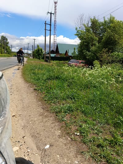 Two cyclists are riding on the edge of a paved road in Andreyevo, Russia. One is in the foreground, and the second is behind him. To the right of the road, a dirt path runs alongside overgrown vegetation. A red car is partially concealed within the foliage near a house with a green roof. Utility poles line the road, and a red and white cell tower is visible in the background. The sky is blue with scattered clouds. The overall scene appears rural or suburban.