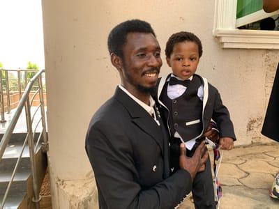 The image depicts an adult male holding a young child in an outdoor setting in Kurudu, Nigeria. The adult male, with dark skin, dark hair, and a beard, is smiling and wearing a black suit jacket over a white collared shirt. He is supporting the young child, who also has dark skin and short dark curly hair, and is dressed in a black suit jacket, black waistcoat, white collared shirt, and a black bow tie. The child, with a neutral expression, holds a colorful patterned strap in their left hand area. Both individuals are positioned in front of a light-colored concrete column and wall. To the left of the column, a silver-colored metal handrail of a staircase is visible, with green foliage and sky in the background. To the right, a window frame with a green element is partially visible above a paved ground area.