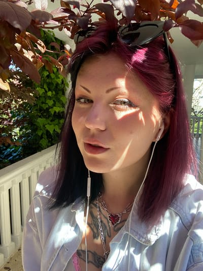 A young woman with dyed burgundy hair is the focal point of the image. Her hair transitions to black at the roots. She is wearing white earphones and has sunglasses perched atop her head. She also has necklaces and visible tattoos on her chest. Her eyes are hazel. She is wearing a light blue denim jacket. She is situated outdoors. In the foreground, there are reddish-brown leaves of a tree or shrub. In the background, there is green foliage and a white railing, which suggests the location may be a balcony or patio. The image is taken in bright sunlight, casting strong shadows across her face. The location of Kunje, Montenegro, cannot be verified from the image.