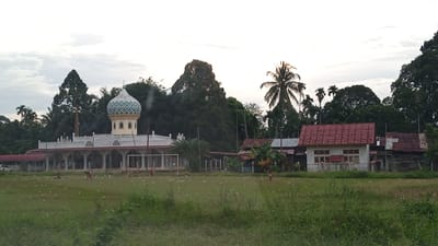 The image shows a mosque with a distinctive turquoise and white dome, flanked by a white building with arched colonnades. In front of the mosque is a grassy field with a soccer goal, scattered with white debris. Three children are visible on the field, one appearing to play soccer. To the right of the mosque are two more buildings, one with a red tiled roof and white walls, and another with a corrugated metal roof. Tall trees, including palm trees, form a dense backdrop. The sky is overcast. The scene is identified as being in the city of Cotmerbo, Indonesia.