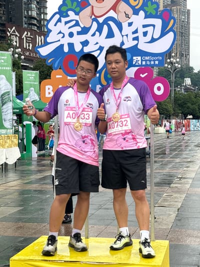 Two men are posing for a photo after an event; they are standing in front of signage from "CMSports 亚马体育," wearing purple and pink shirts with numbers "A0732" and "A0/33" pinned to them and gold medals around their necks. They are giving a thumbs up. The ground is paved, and in the background is a partially visible building with a sign saying "SICHUA." To the left are banners and items that appear to be water bottles and paper towels, and the edge of a yellow object. The city is Nan'an District, China.