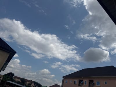 A bright daytime shot of a cloudy sky over buildings in Enugu, Nigeria. The sky is a clear blue with scattered white cumulus clouds, some of which are quite large and billowy. On the left side of the image, the dark tiled roof and light-colored wall of a building are visible, angled towards the sky. In the lower right quadrant, another building with a dark tiled roof and a light pink facade is prominent, featuring multiple windows and balconies. Further in the distance, other buildings and trees can be seen under the expansive sky. The overall scene suggests a typical day in a residential or urban area.