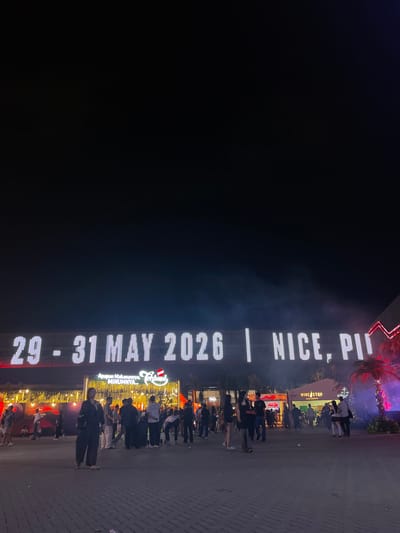 The image shows a night scene in Salembaran, Indonesia, where a crowd of people gathers near an illuminated vertical sign reading "29-31 MAY 2026 | NICE, PI." The sign also features an advertisement for a restaurant or food-related establishment with the text "Apapun Makanannya, MINUMNYA" and the name "Tenbord." A "WINE STOP WINE & SPIRITS" storefront is visible behind the crowd. Palm trees adorned with lights add to the nighttime ambiance. The foreground shows paving stones.