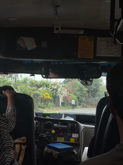 This image captures the interior perspective from within a public transit bus in El Congo, El Salvador, looking forward through the windshield. In the immediate foreground, the backs of two passengers' heads and shoulders are visible on the left and right; the person on the left wears a black and white striped shirt with their right arm raised. Directly in front, the bus's dashboard is visible, featuring various controls, vents, and a radio. A black cup in a holder and a blue rectangular box rest on the dashboard. Overhead, a rearview mirror is centrally positioned, reflecting a partial face. A small, black electric fan hangs from the mirror's support structure. Above the windshield, a partial white sign reads "...ERS MUST NOT STAND FORWARD OF WHITE LINE WHILE BUS IS IN MOTION," and a yellow sign labeled "BUS SAFETY RULES" is affixed to the dashboard panel. To its right, a white document displays "AB 248," "$0.70," and "URBANO," indicating a fare for urban transport.

Through the windshield, a paved road with a yellow centerline extends forward. The exterior environment is lush and verdant, characterized by abundant tropical vegetation including trees, shrubs, and numerous flowering plants with white, yellow, pink, and red blossoms lining the roadside. On the left side of the road, a male individual wearing a white shirt and dark pants is visible walking away from the bus. The sky appears overcast or hazy.