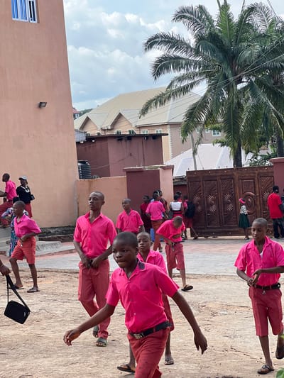 The image depicts a group of young people, predominantly boys wearing matching pink shirts and red shorts, in an outdoor setting. The architecture suggests an urban or semi-urban environment, possibly a school or a community center, with a prominent building featuring a light-colored stucco facade and a blue-paned window on the left. In the background, other buildings with beige roofs and some palm trees are visible. A large, ornate wooden gate stands on the right side of the image. Some individuals are in motion, walking or looking around, indicating a gathering or a school activity. The ground appears to be paved or a dirt-like surface. The overall scene is brightly lit by daylight under a cloudy sky. It is highly probable that this image was taken in the city of Nnewi, Nigeria, given the context of typical school attire and architectural styles found in the region.