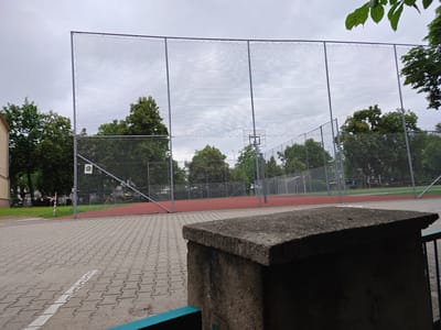 The image shows an outdoor sports facility with a tall chain-link fence enclosing a red track and a green field. The sky is overcast and cloudy. In the foreground, there is a low concrete structure, possibly a bench or a planter, with a teal-colored railing to its right. The ground is paved with interlocking concrete bricks. Trees surround the sports area, and a building with a beige facade is visible on the left. The scene appears to be in a public park or schoolyard. The location is identified as the "Orlik" sports field at ul. Wileńska 46 in Poznań, Poland.
